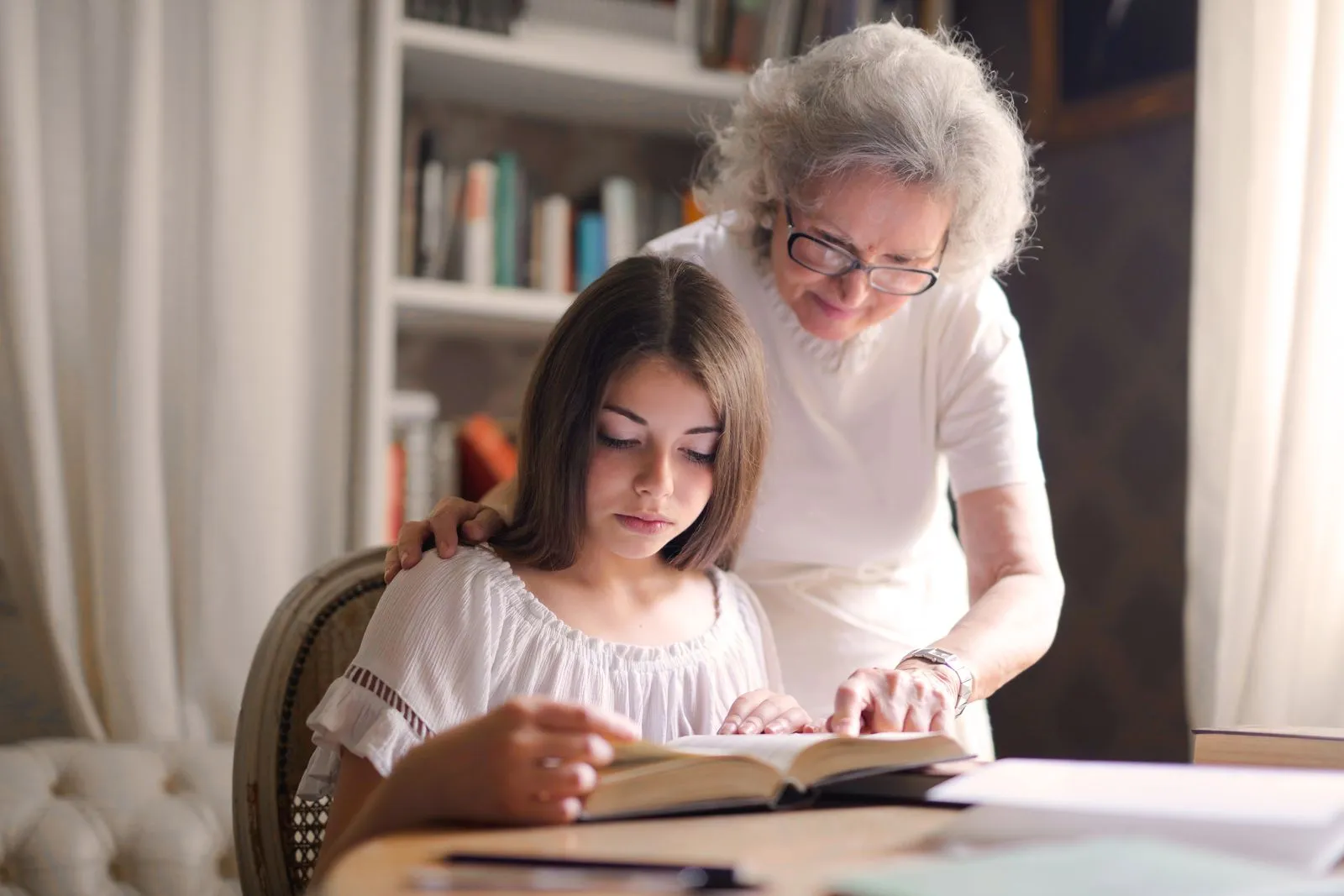 A grandmother helping her granddaughter read a book at a desk